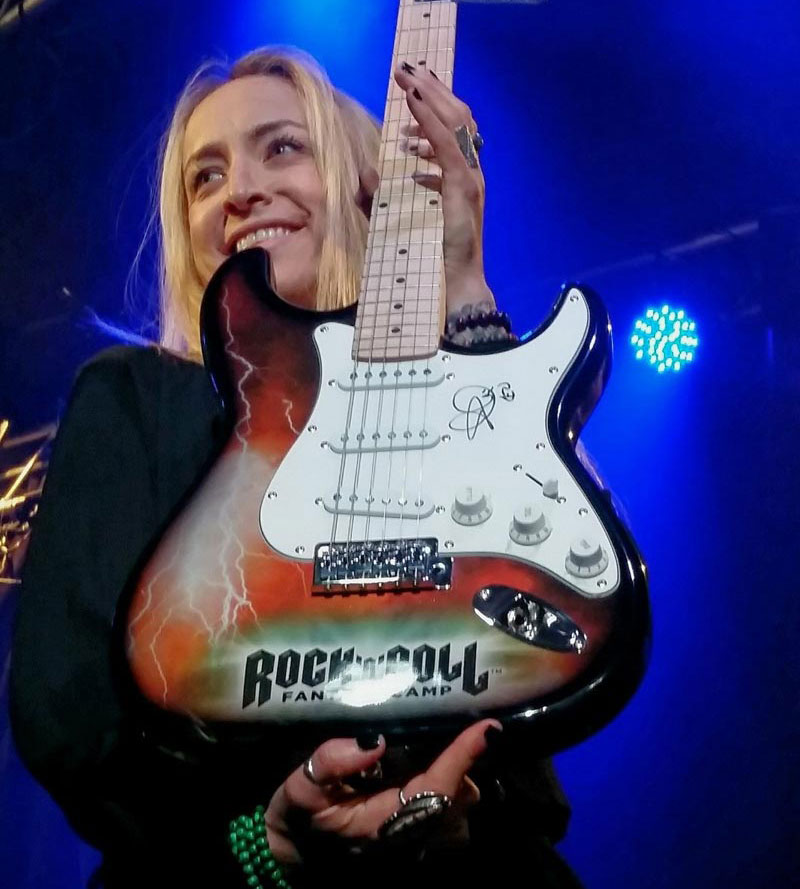 Woman on stage, holding a custom, autographed Rockcamp guitar